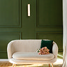 A tall matt gold wall light with a glowing wave‑ridged glass diffuser mounted on a dark green panelled wall above a cream upholstered sofa with a green cushion and a bouquet of flowers.