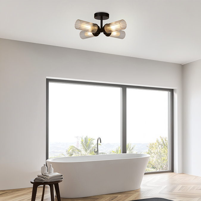 A matt black four‑light flush ceiling fixture with ribbed glass shades mounted in a modern bathroom above a freestanding bath, softly illuminating a bright space with large windows.