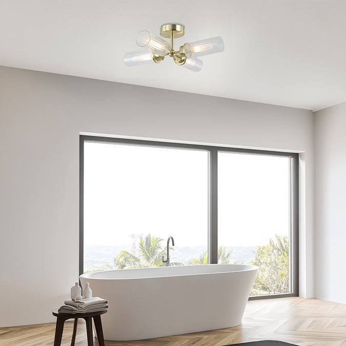 A satin brass four‑light flush ceiling fixture with ribbed glass shades mounted in a modern bathroom above a freestanding bath, softly illuminating a bright space with large windows.