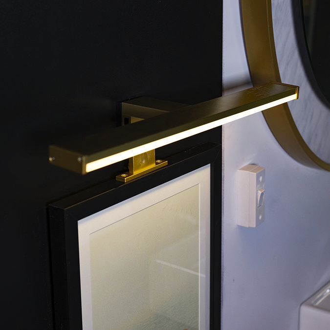 A slim matt‑gold LED mirror light mounted above a small rectangular mirror, casting warm illumination against a dark wall beside a round mirror with a gold rim.