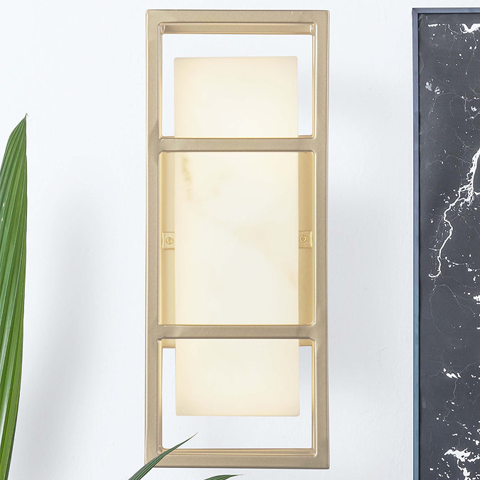 A matt gold rectangular wall light with a glowing marble‑effect resin diffuser mounted on a white wall, shown beside a black marble feature panel and a leafy green plant.