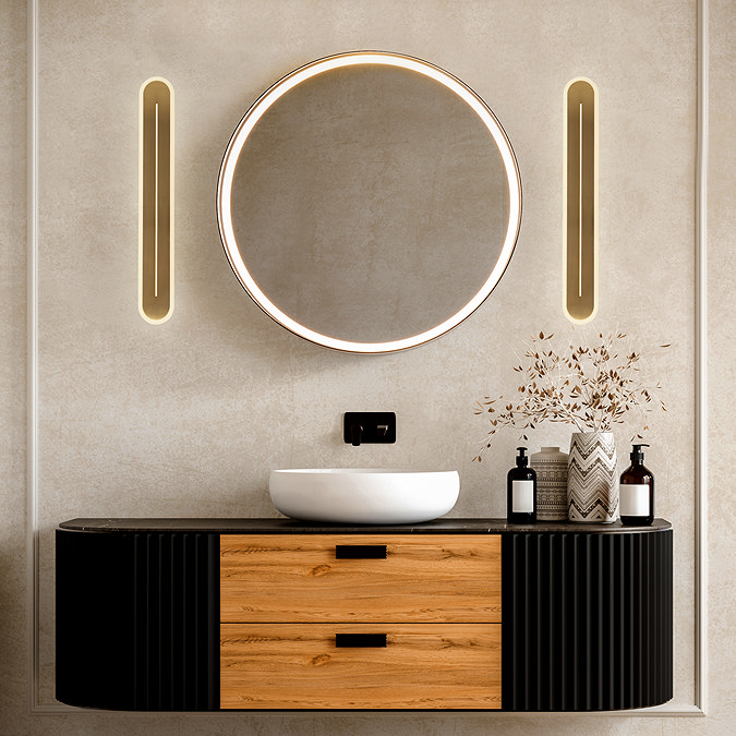 Two tall satin‑brass wall lights with glowing central apertures mounted on either side of a round mirror above a modern bathroom vanity.