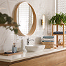 A tall gold clamp‑style wall light with a glowing ribbed opal glass tube mounted on a patterned white bathroom wall beside a round wooden mirror and a wooden shelving unit with folded towels and a plant.
