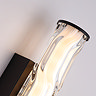 Close‑up of the wall light showing the curved wave‑ridged glass diffuser, the matt black backplate, and the matt black end cap illuminated from within.