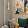 A tall matt black wall light with a glowing wave‑ridged glass diffuser and pull‑cord mounted on a neutral wall beside geometric artwork above a sofa with grey cushions and green and yellow accent pillows.