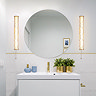 Two tall matt gold wall lights with glowing wave‑ridged glass diffusers and pull‑cords mounted on either side of a large round bathroom mirror above a white vanity with gold accents and decorative items.