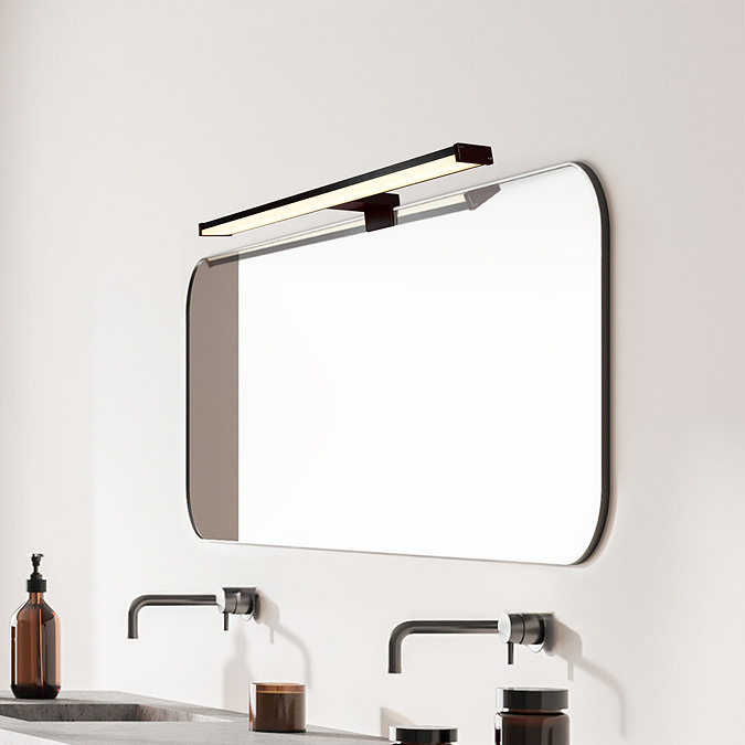A long matt‑black LED mirror light mounted above a wide rectangular bathroom mirror, illuminating a minimalist vanity with wall‑mounted taps and soap dispensers.