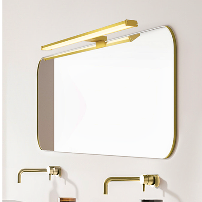A long matt gold LED mirror light mounted above a wide rectangular bathroom mirror, illuminating a minimalist vanity with wall‑mounted taps and soap dispensers.