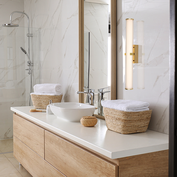 A tall gold clamp‑style wall light with a glowing ribbed opal glass tube mounted beside a large mirror above a modern bathroom vanity with wooden drawers, wicker baskets, and marble‑effect wall tiles.