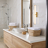 A tall gold clamp‑style wall light with a glowing ribbed opal glass tube mounted beside a large mirror above a modern bathroom vanity with wooden drawers, wicker baskets, and marble‑effect wall tiles.