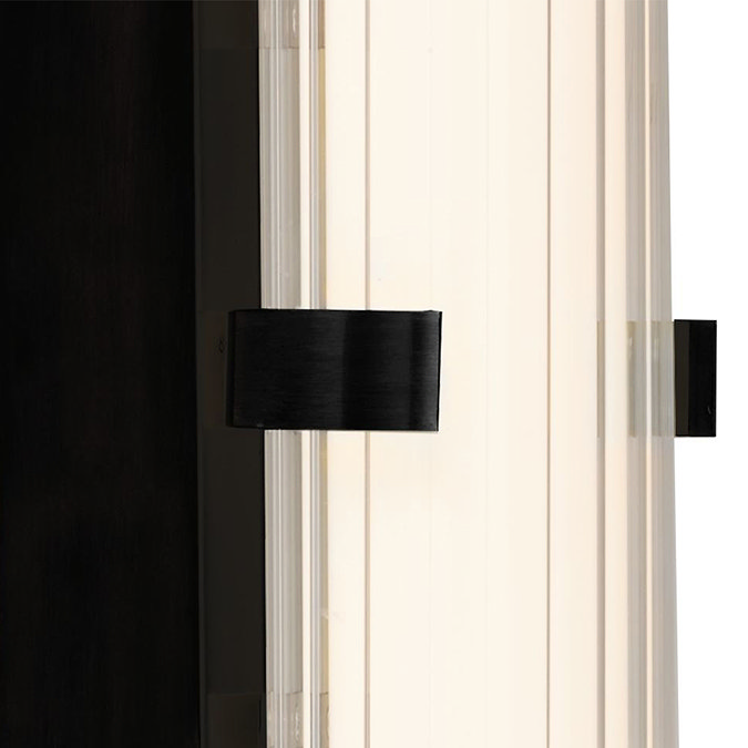 Close‑up of the matt black clamp wall light showing the glowing ribbed opal glass tube and the black metal clamp securing it at the centre.