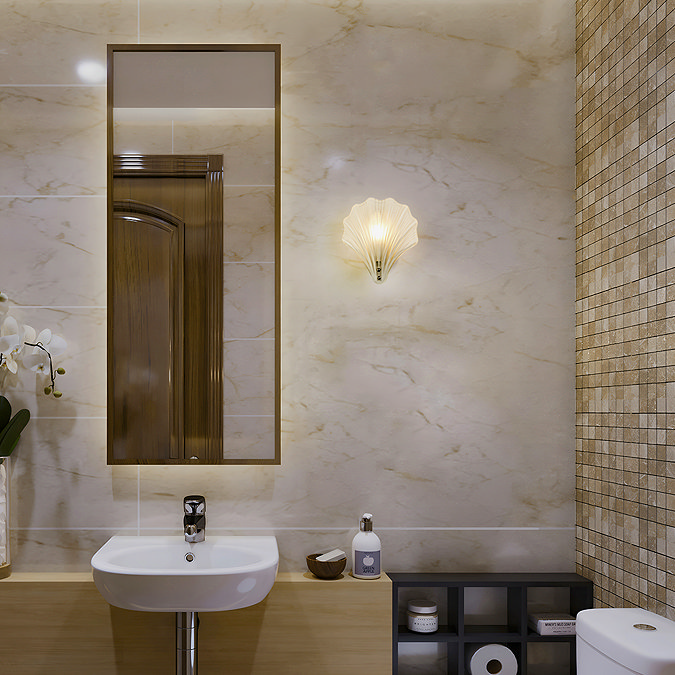A frosted scallop‑shaped wall light with a satin silver fitting mounted on a marble‑effect bathroom wall, casting a soft warm glow beside a sink and a tall mirror.