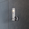 A chrome bathroom wall light with a clear etched glass shade mounted on a dark tiled wall, shown illuminated.