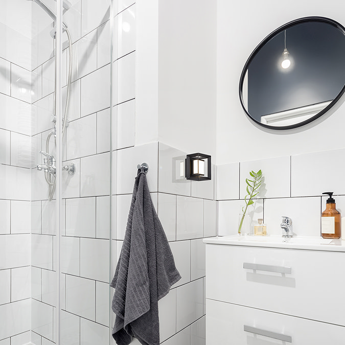 A small black square wall light with an opal diffuser mounted beside a bathroom sink and round mirror, illuminating white tiled walls near a glass shower enclosure.