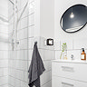 A small black square wall light with an opal diffuser mounted beside a bathroom sink and round mirror, illuminating white tiled walls near a glass shower enclosure.