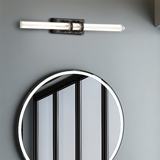 A slim matt black wall light with a glowing glass diffuser mounted above a large round mirror on a grey bathroom wall.