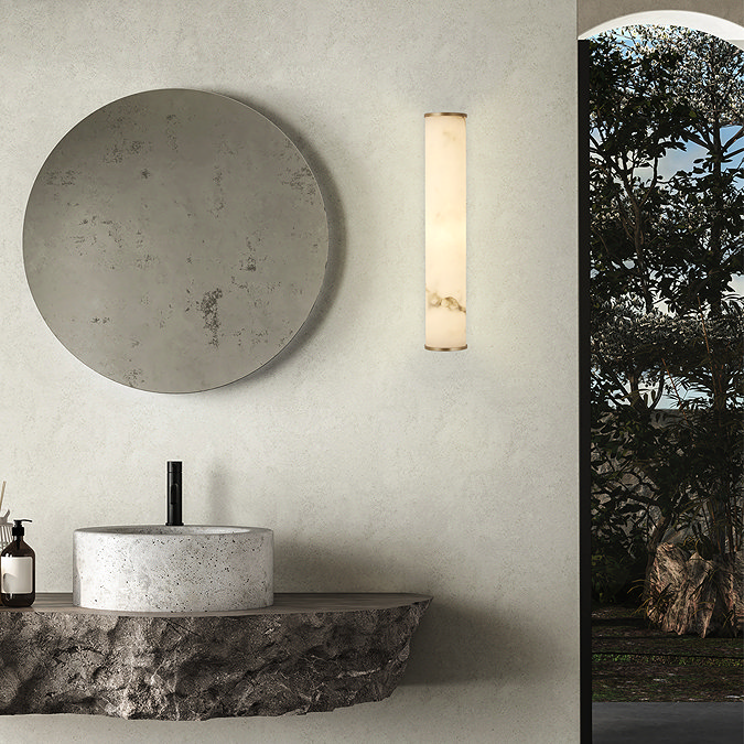 A tall matt‑gold wall light with a glowing marble‑resin diffuser mounted beside a large round mirror above a stone basin in a minimalist bathroom with textured walls.