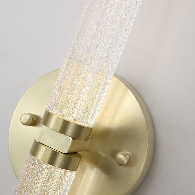 A close‑up view of the satin‑brass wall light’s base. The image highlights the brushed brass finish, the mounting screws, and the detailed texture of the clear acrylic light tube that attaches to the centre of the round base.