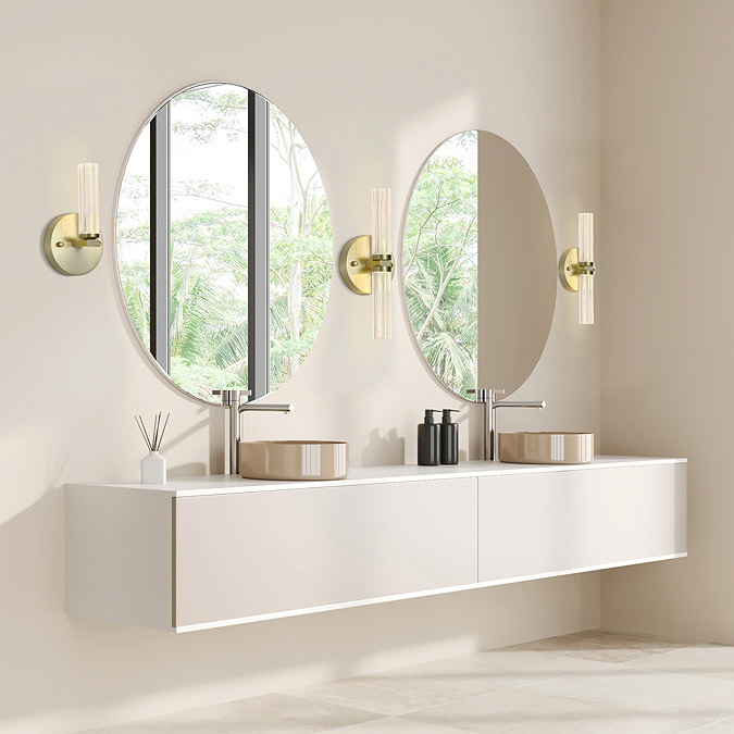 A bright, modern bathroom with two round mirrors mounted above a long floating vanity unit. On each side of both mirrors is a matching satin‑brass wall light with textured acrylic cylinders, creating symmetrical and soft illumination. The room features neutral colours, large windows, and minimalist décor.