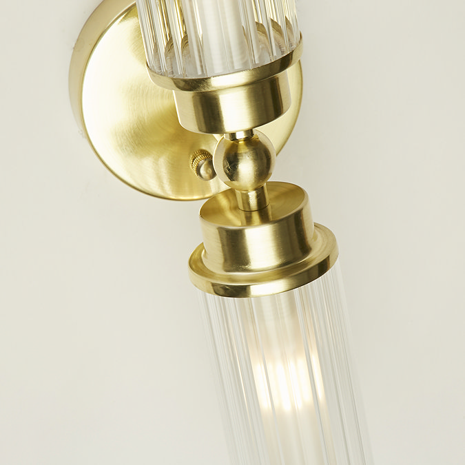 Close‑up of the satin brass up‑and‑down wall light showing the ribbed glass shades and rounded brass detailing, with warm light glowing inside the lower shade.