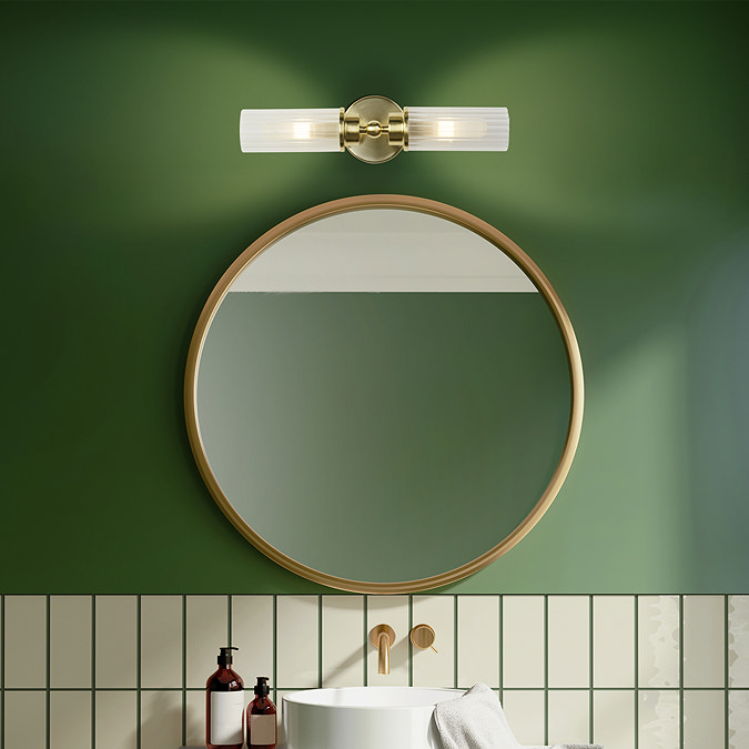 A satin brass up‑and‑down wall light with two ribbed glass shades mounted above a round mirror in a bathroom with green and tiled walls, casting a warm glow over the basin area.