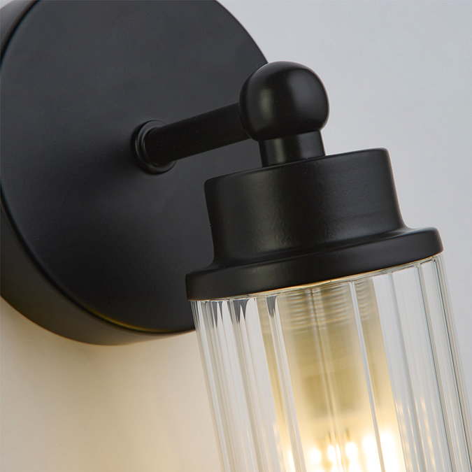 Close‑up of the matt black fitting and ribbed clear glass shade of the wall light, showing soft illumination inside the glass.