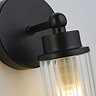 Close‑up of the matt black fitting and ribbed clear glass shade of the wall light, showing soft illumination inside the glass.