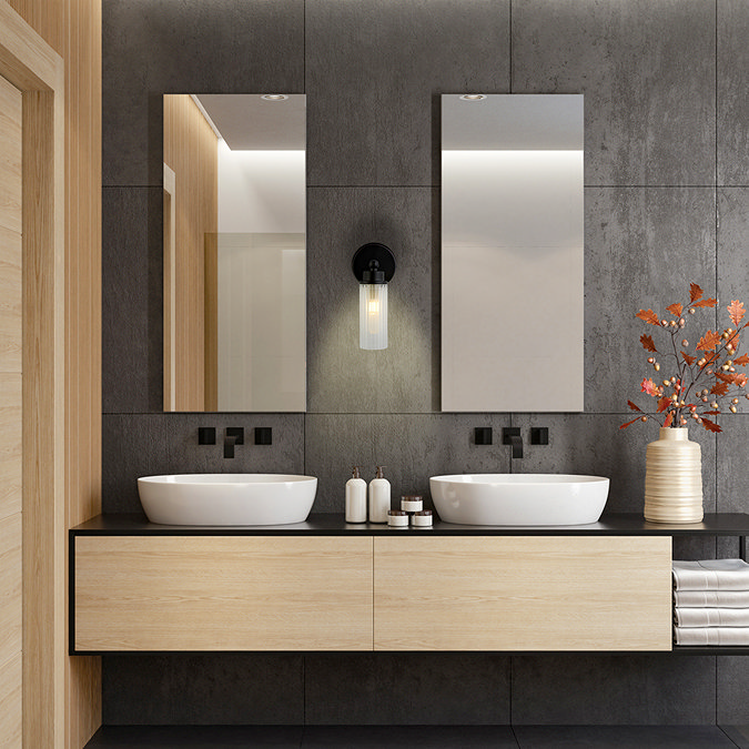 A matt black wall light with a clear ribbed glass shade mounted between two bathroom mirrors on a dark tiled wall, softly illuminating a twin‑basin vanity area.