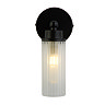 A matt black wall light with a clear ribbed glass cylinder glowing warmly, supported by a round black wall plate.