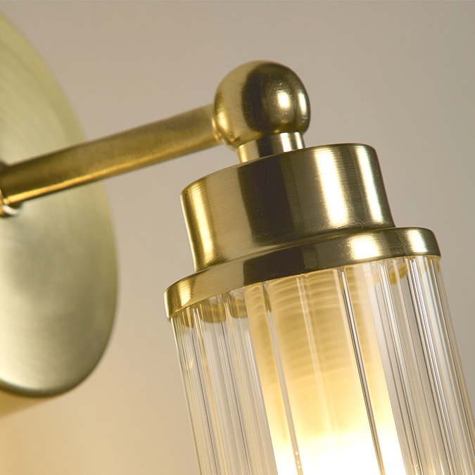 Close‑up of the satin brass fitting and ribbed clear glass shade of the wall light, showing soft illumination inside the glass.