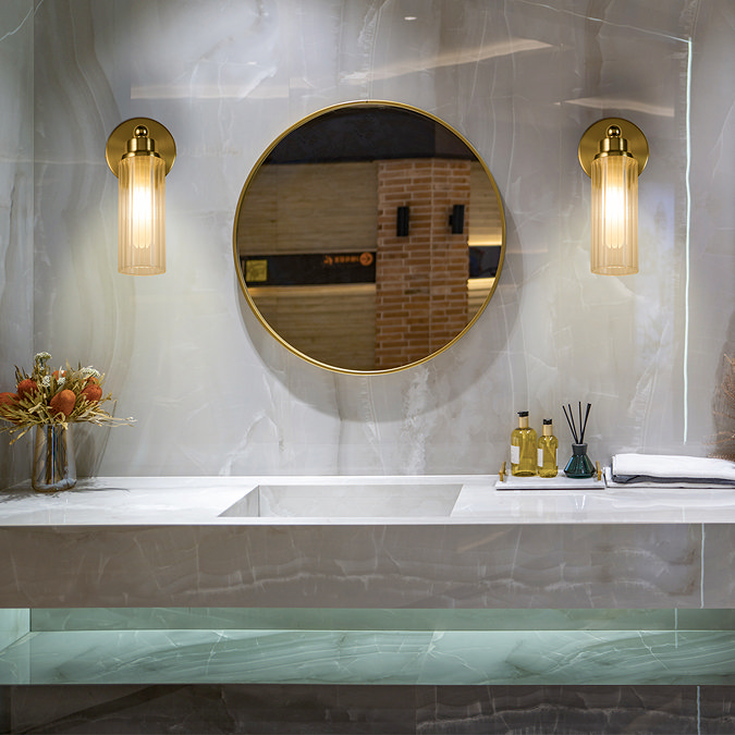 Two satin brass wall lights with clear ribbed glass shades mounted beside a round mirror above a marble bathroom vanity, casting a warm glow across the space.