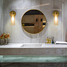 Two satin brass wall lights with clear ribbed glass shades mounted beside a round mirror above a marble bathroom vanity, casting a warm glow across the space.