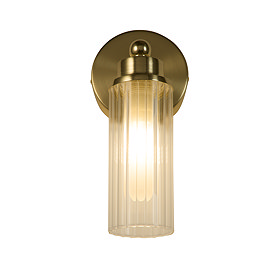A satin brass wall light with a clear ribbed glass cylinder glowing warmly, supported by a round brass wall plate.